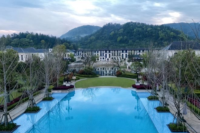 A large rectangular pool with a clear blue surface, surrounded by a landscaped area with trees and a view of a building complex in the background.