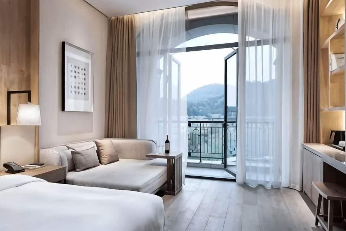 A hotel room with two beds, a desk, a chair, a lamp, a framed picture, and a balcony with a view.