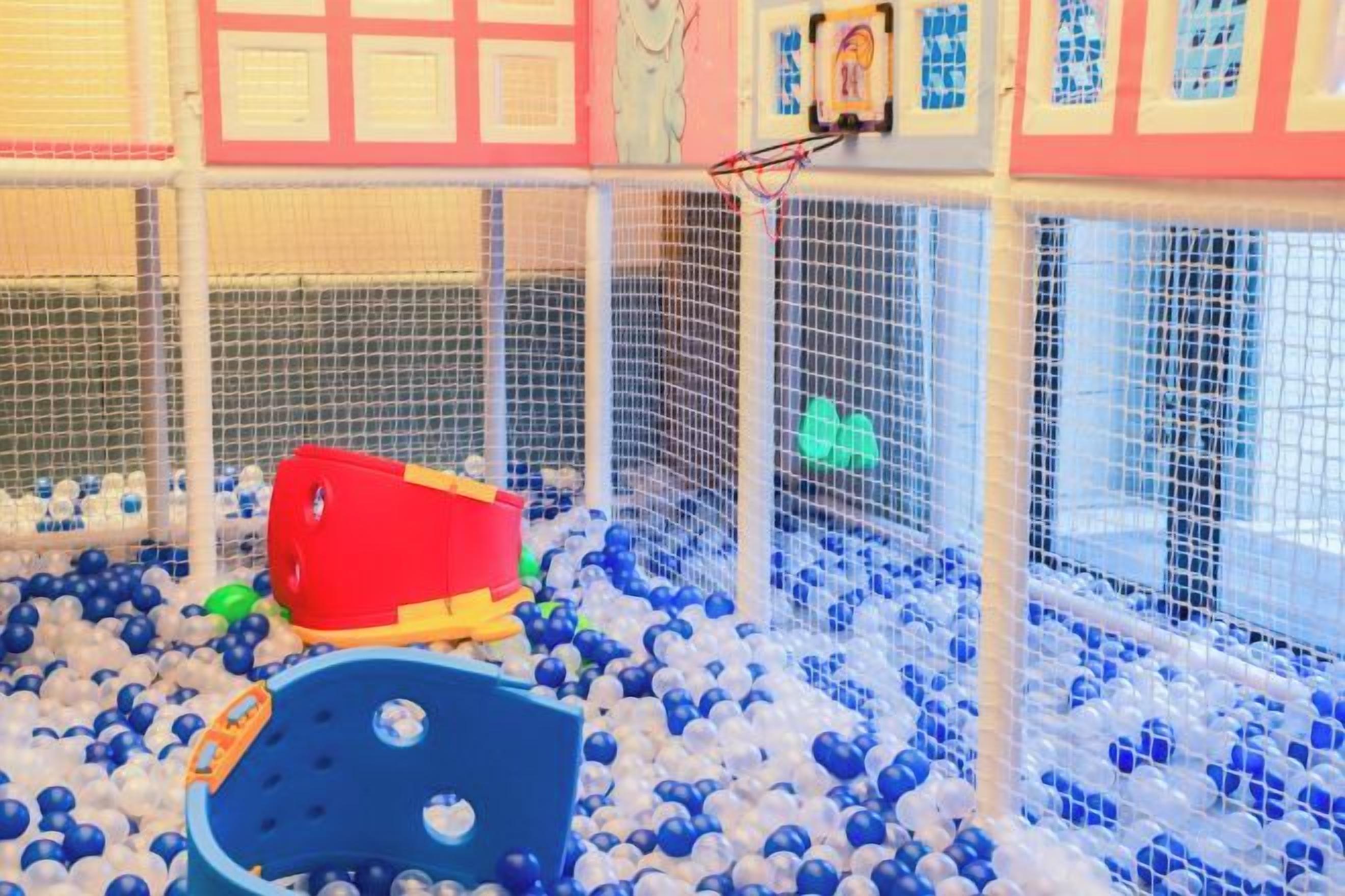 Un área de juegos con piscina de pelotas, un módulo de juegos rojo y amarillo, y un aro de baloncesto.