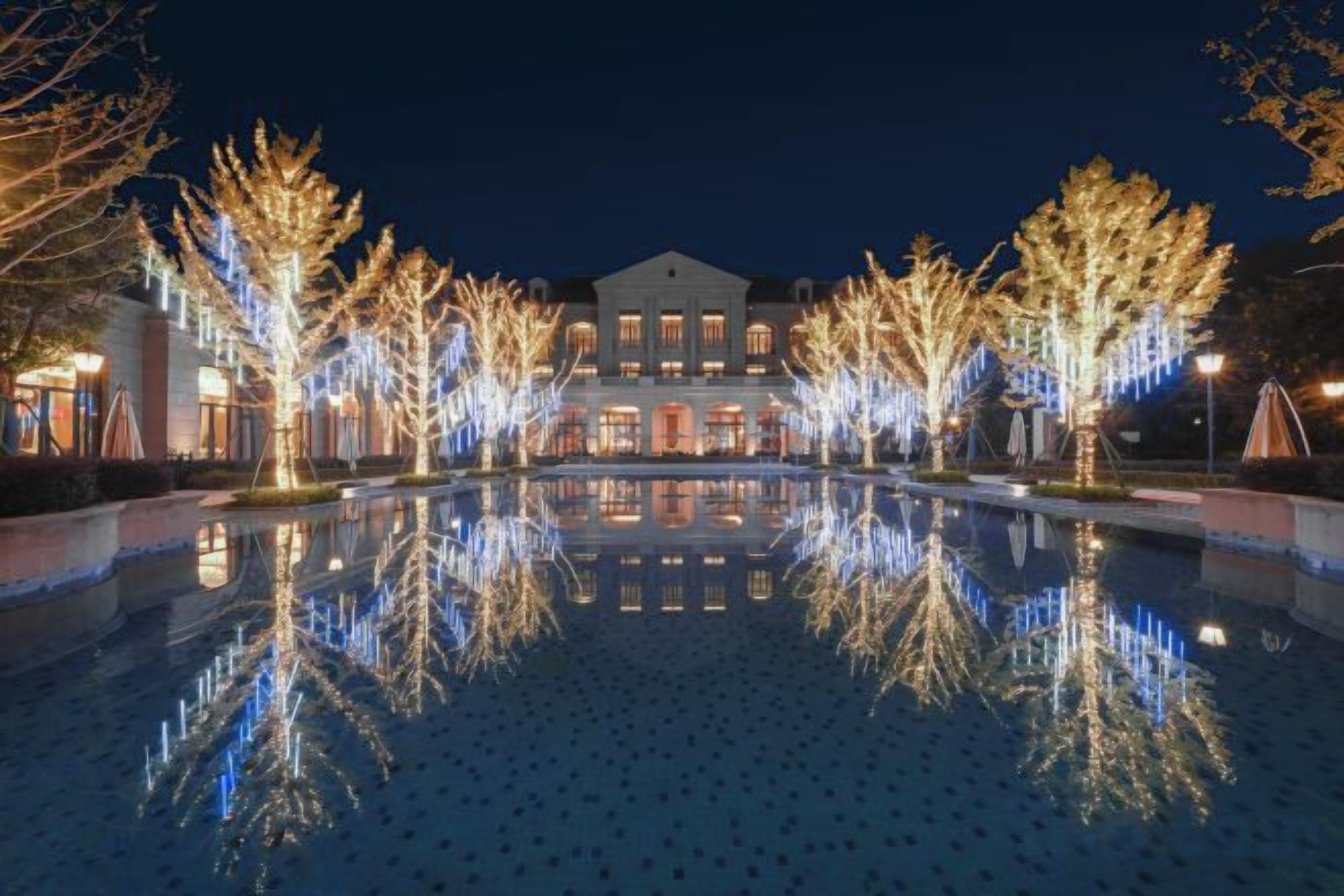 A nighttime view of a hotel with a reflective pool, trees with lights, and a building with multiple windows.