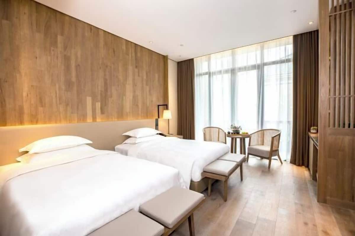 A hotel room with two beds, a small table, a chair, and a large window.