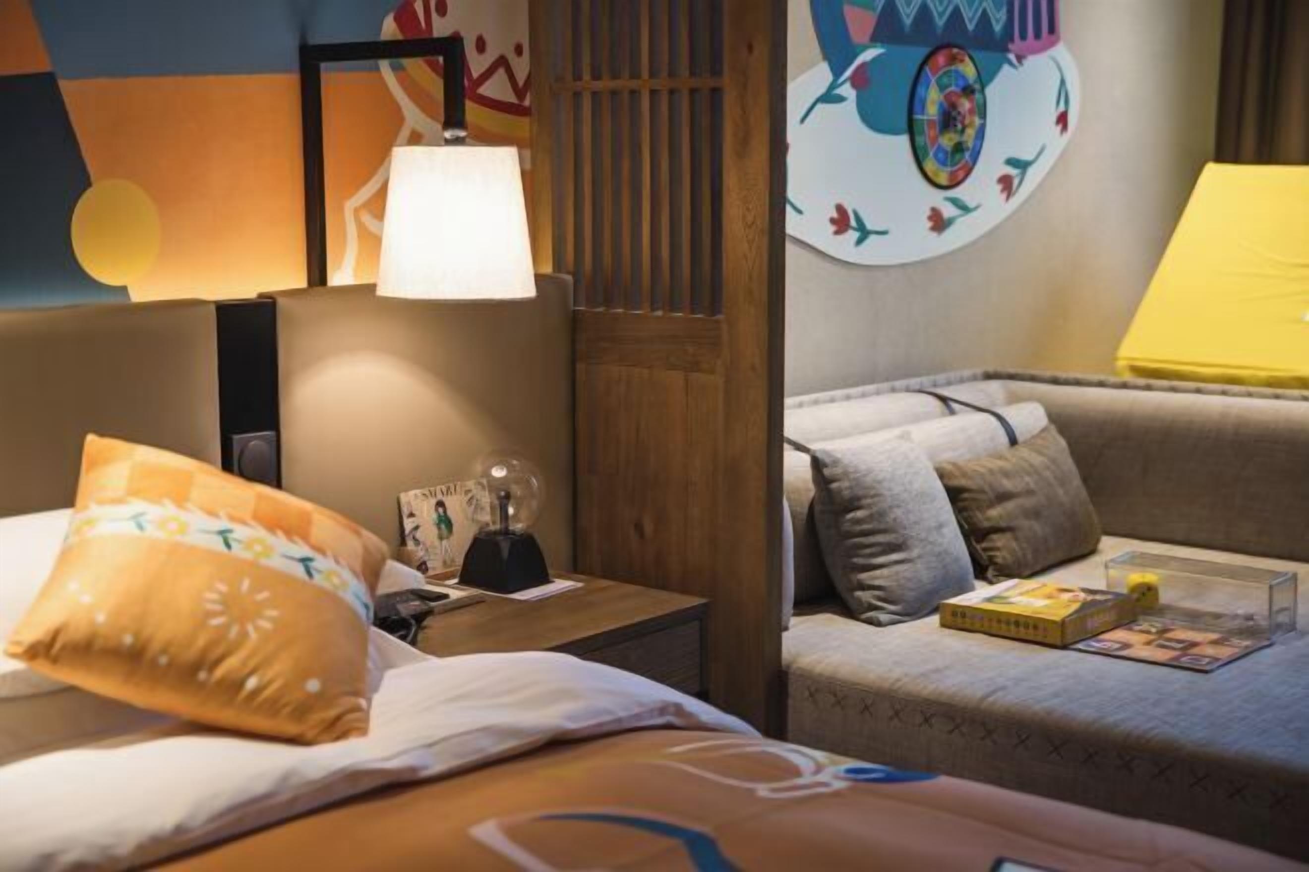 Habitación de hotel con cama, mesita de noche, lámpara y una pared decorada con una obra de arte colorida.