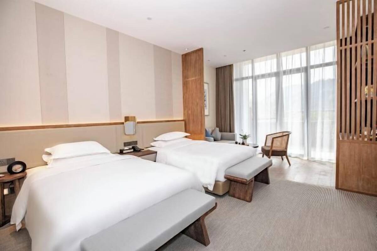 A hotel room with two beds, a bench, a desk, a chair, and large windows.