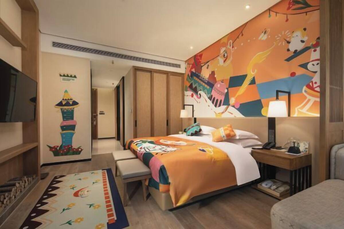 A hotel room with a bed, bedside table, lamp, and a colorful mural on the wall.