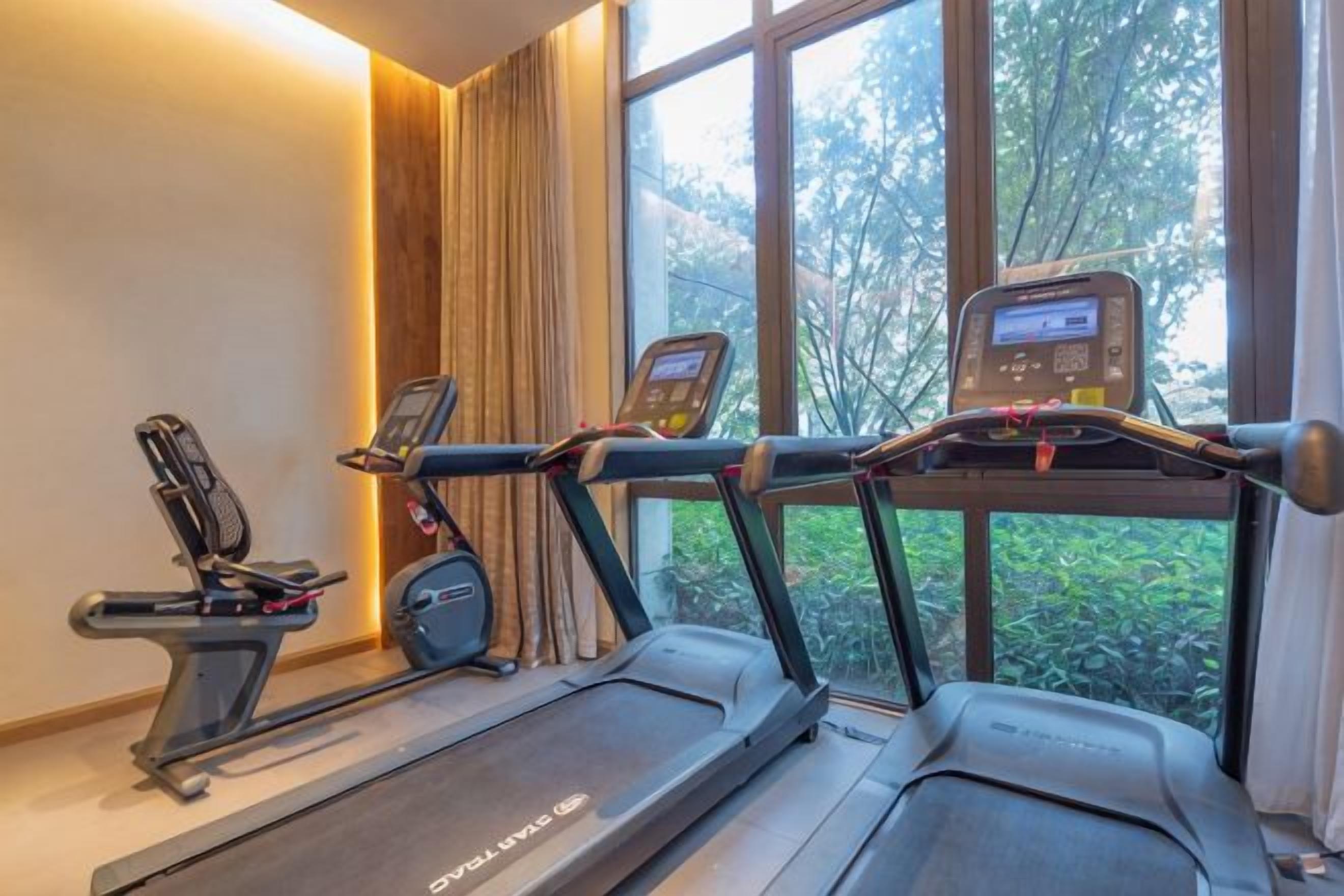 A fitness area with treadmills and an elliptical machine, a digital display, and a window with a view of trees.