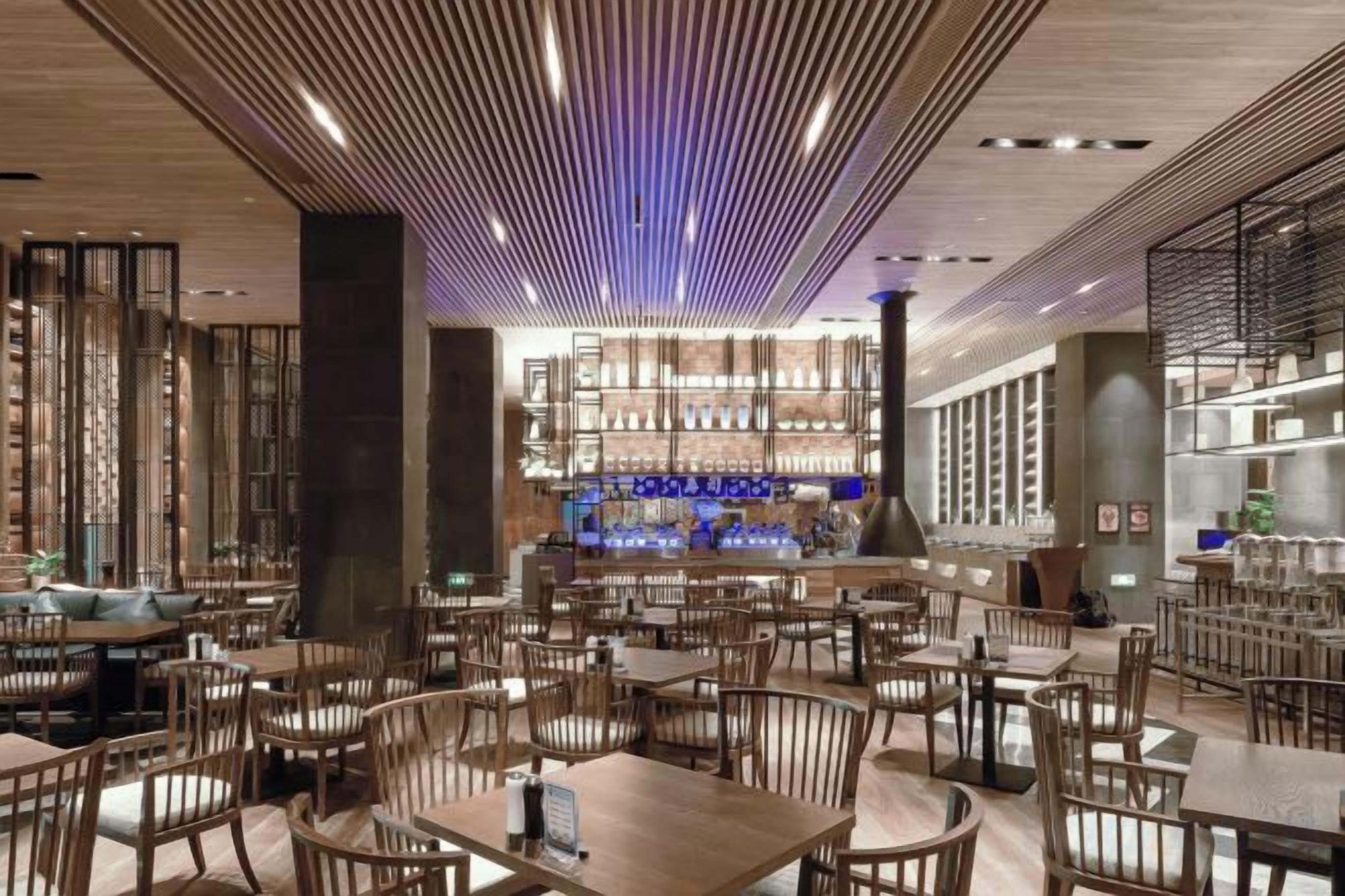 Un restaurante con varias mesas y sillas, una zona de bar al fondo y un diseño moderno en el techo.