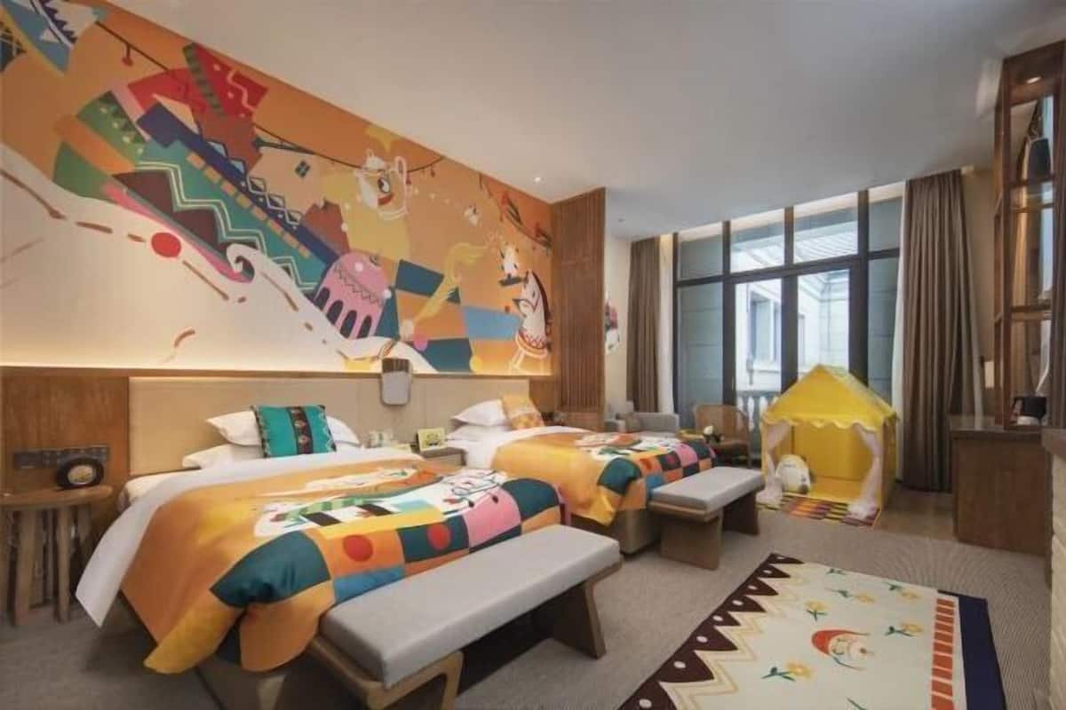 A hotel room with two beds, a large mural, a window with curtains, a small playhouse, and a desk with a lamp.