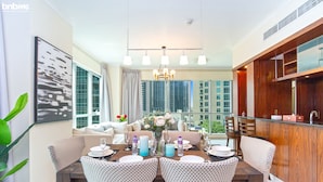 A dining area with a wooden table set for four, surrounded by patterned chairs. A chandelier hangs above, and a large window offers a view of high-rise buildings. - bnbme | The Residences | 2 BDR (Dubai)