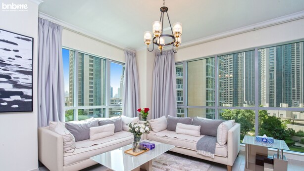 A spacious living room with a large sectional sofa, a glass coffee table, and a view of tall buildings through large windows. - bnbme | The Residences | 2 BDR (Dubai)