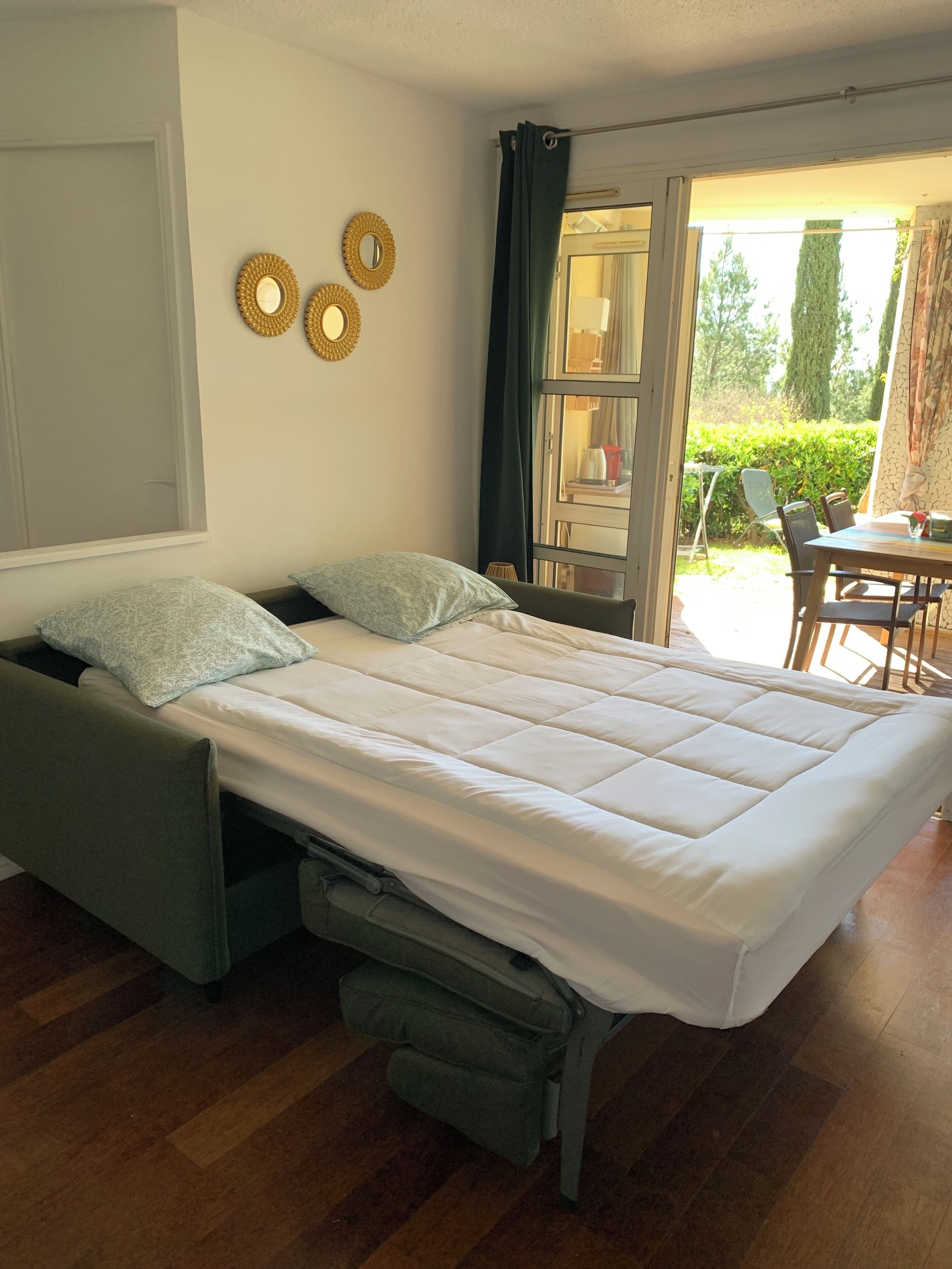 Ein Zimmer mit einem Bett, einem kleinen Tisch und einem Stuhl. Drei Spiegel sind an den Wänden angebracht und es gibt ein Fenster mit Vorhängen.