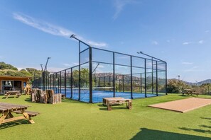 Een tennisbaan buiten met een net, omringd door kunstgras, picknicktafels en een klein gebouw met een barbecueplaats.
