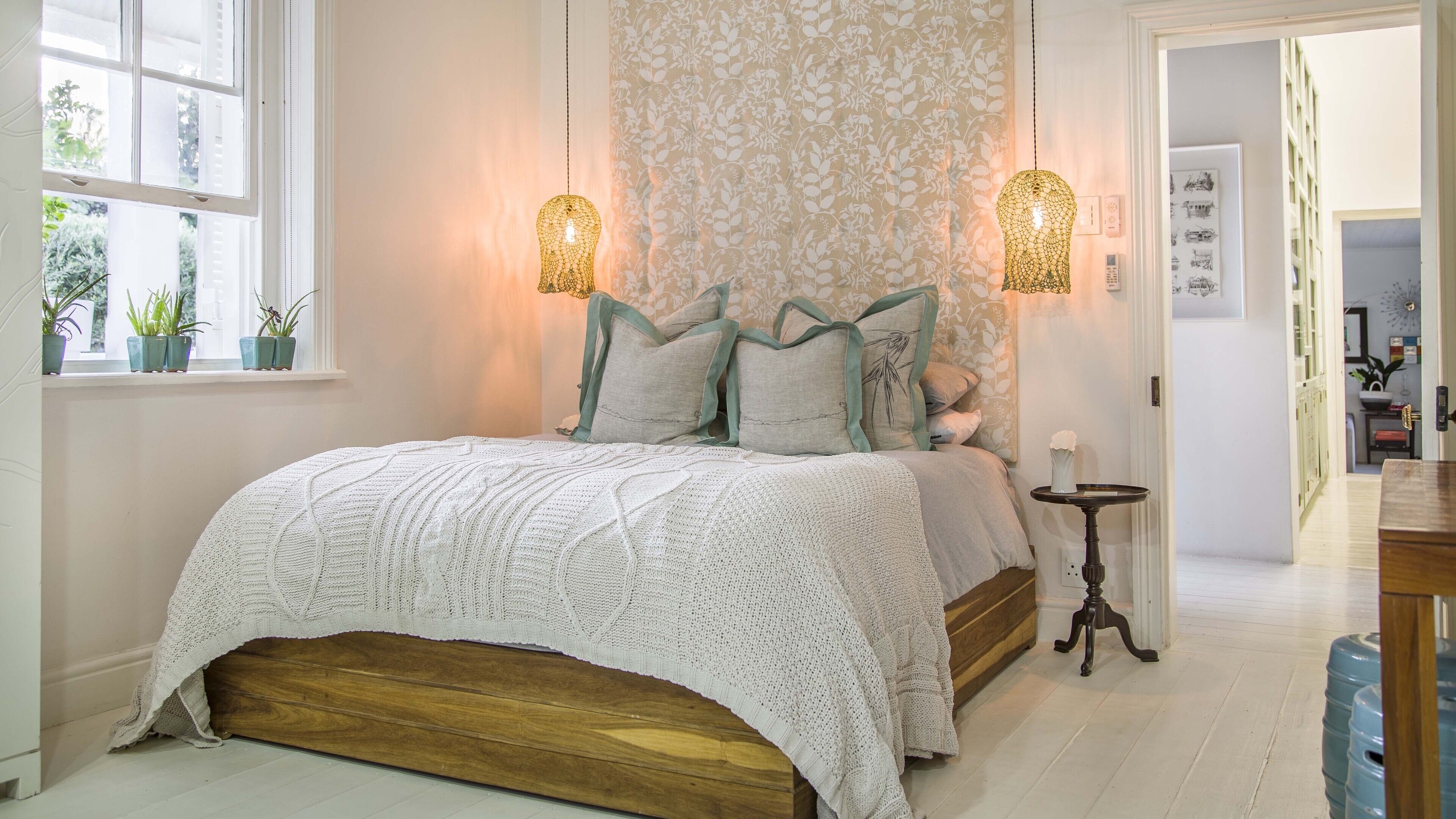 A bedroom with a wooden bed, white bedding, and a floral-patterned wall. There are two wall-mounted lamps and a small table with a vase.
