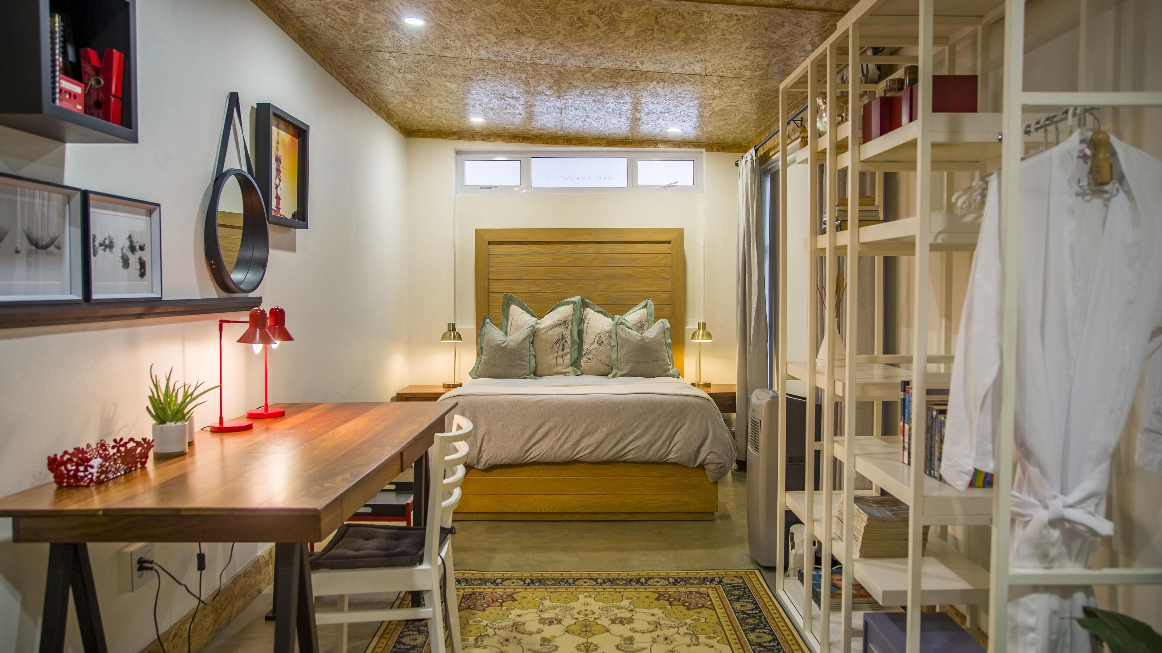 A bedroom with a wooden headboard, a bed with pillows, a desk with a lamp, a wardrobe, and a small plant.