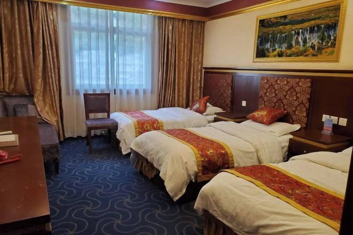 A hotel room with two beds, a desk, and a chair. There is a painting on the wall and a window with curtains.