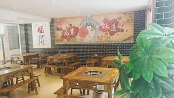 Interior de restaurante com mesas e cadeiras de madeira, uma parede decorativa grande e um vaso de planta.