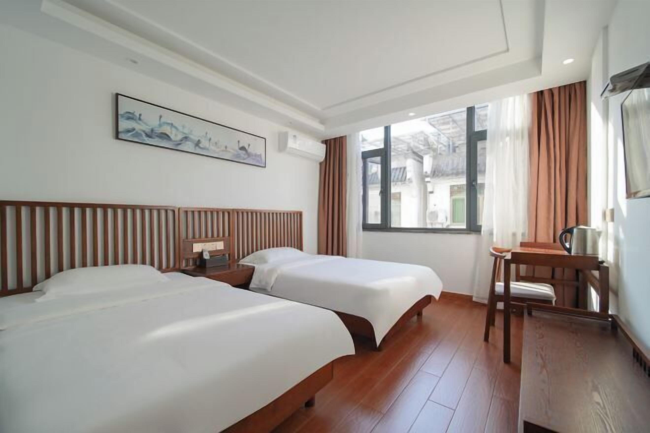 Une chambre d’hôtel avec deux lits, une tête de lit en bois, un bureau avec une chaise, une fenêtre avec des rideaux et un tableau accroché au mur.