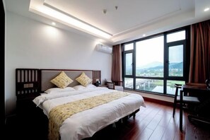 Ein Hotelzimmer mit einem großen Bett, einem Schreibtisch und Blick auf eine Stadtlandschaft durch große Fenster.