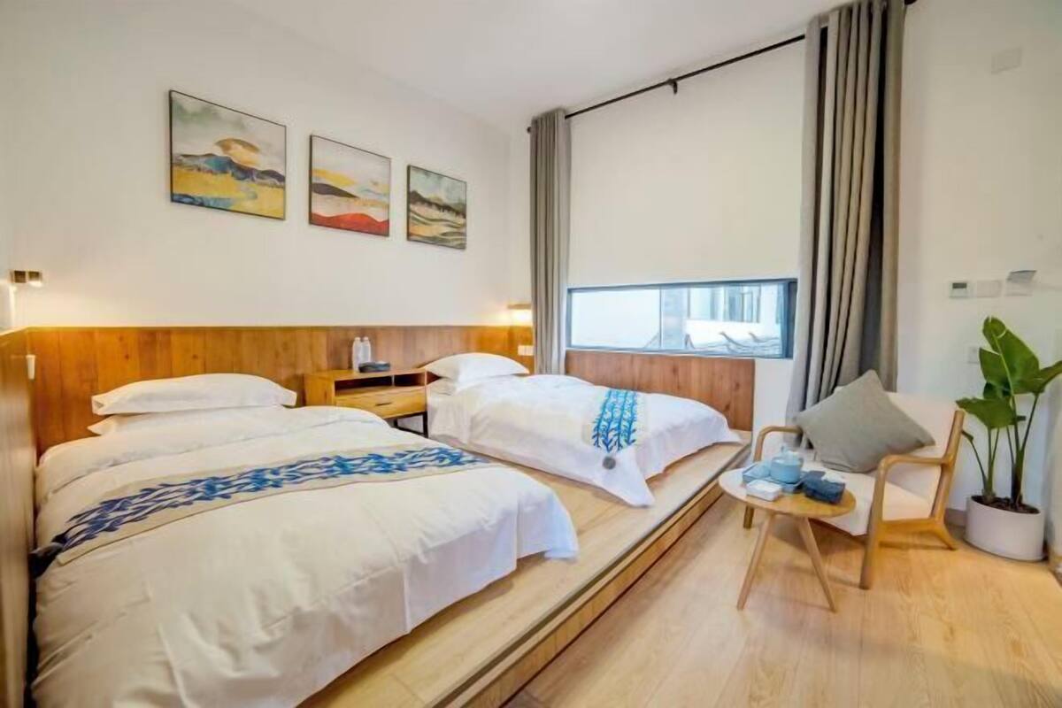 A hotel room with two beds, a wooden headboard, a window with curtains, a small table with a plant, and artwork on the wall.