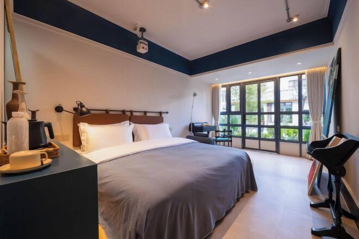 A hotel room with a large bed, a desk with a chair, a window with curtains, and a ceiling light.