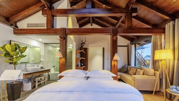 A modern hotel room with a large bed, wooden beams, a sofa, a desk, and a bathroom with a mirror and sink. - Amila Guesthouse (Dali City)