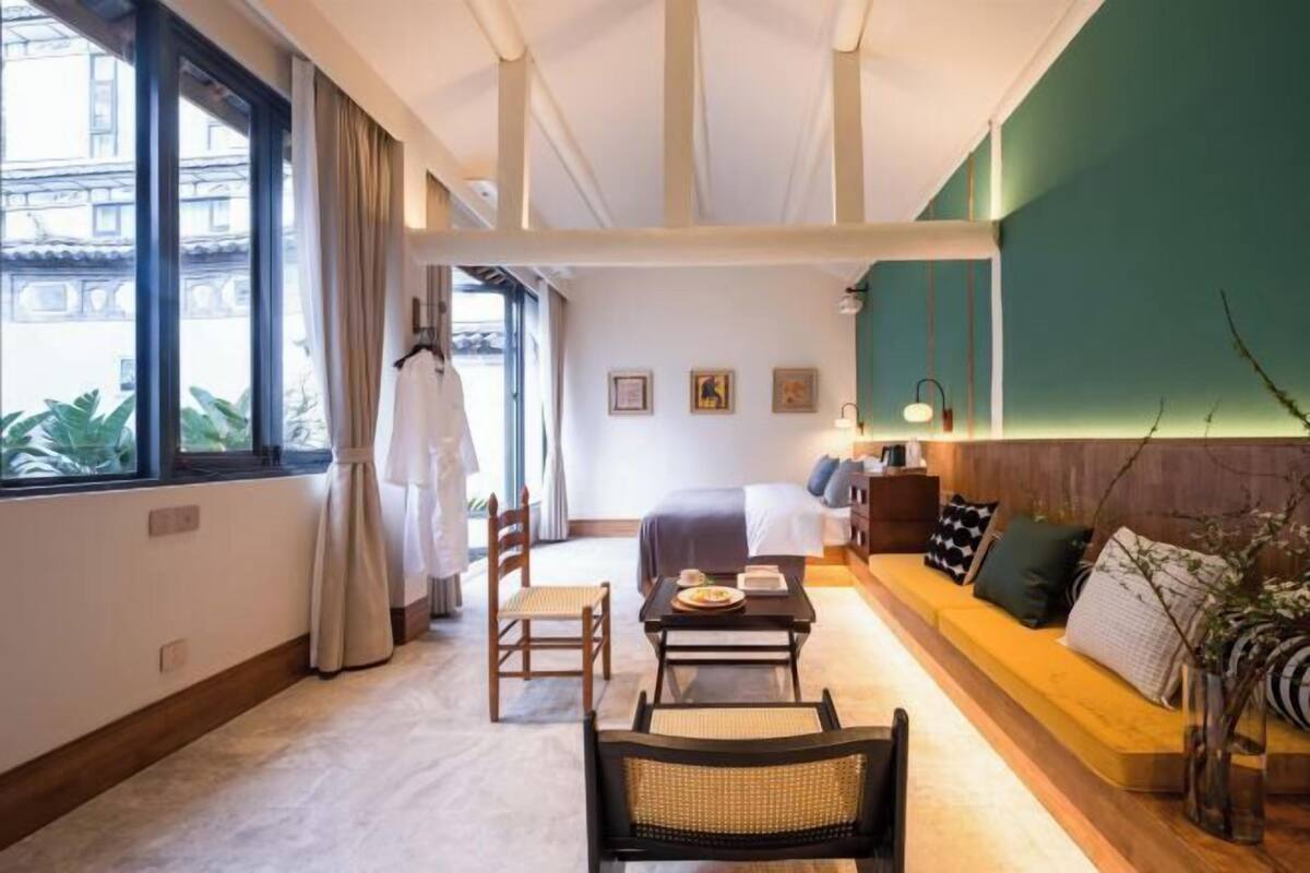 A hotel room with a bed, a sofa, a dining table, and chairs. There is a window with curtains, a wall-mounted lamp, and a plant.