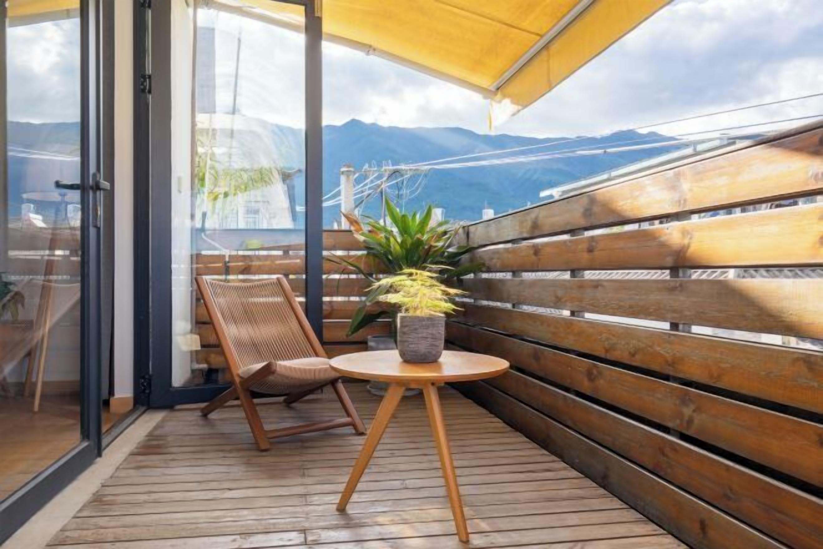 A balcony with wooden flooring, a round wooden table, a wicker chair, and a potted plant.