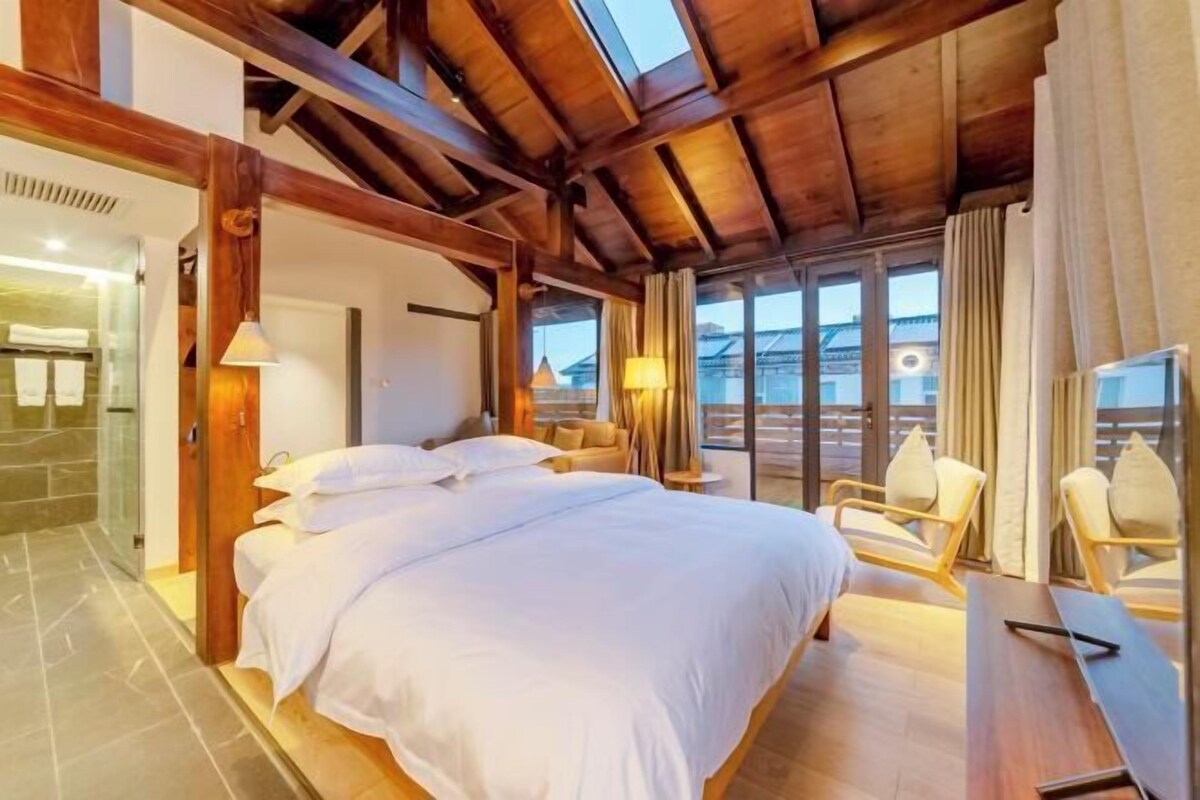 A bedroom with a large bed, a sitting area with chairs, a wooden ceiling, and a view through large windows.