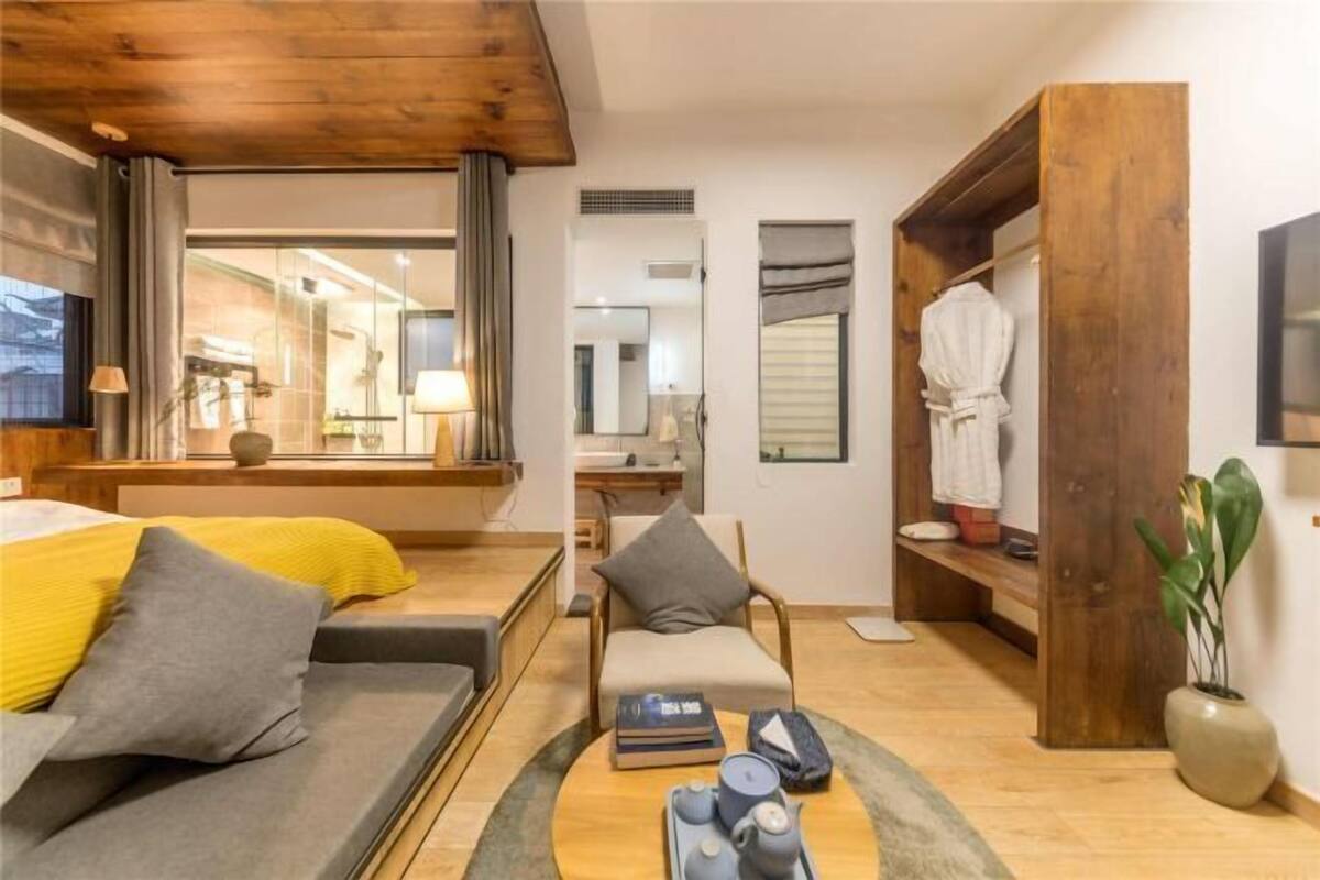 A modern hotel room with a wooden ceiling, a sofa with cushions, a coffee table with books and a teapot, a robe hanging on a wooden rack, and a plant.