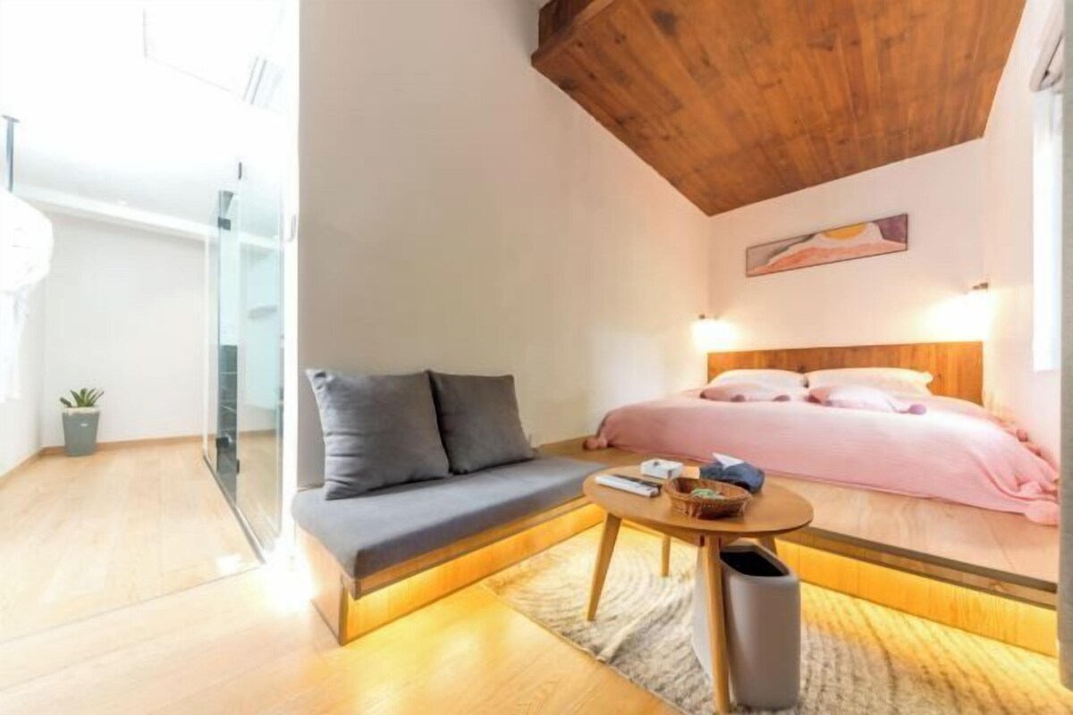 A modern room with a wooden ceiling, a bed with pink bedding, a gray sofa, a small round table, and a glass partition.