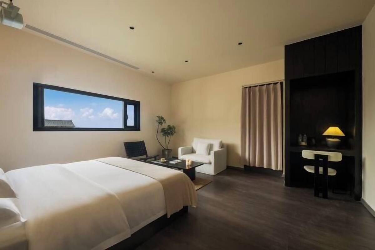 A hotel room with a bed, a desk, a chair, a lamp, and a window with a view.