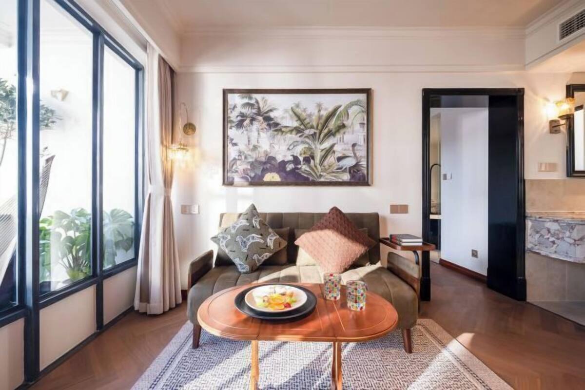 A living room with a sofa, a coffee table with a plate of fruit, a painting on the wall, and a large window.