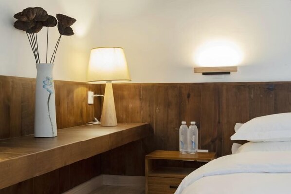 A hotel room with a wooden headboard, a bedside table with a lamp, a vase with dried flowers, and a bed with pillows.