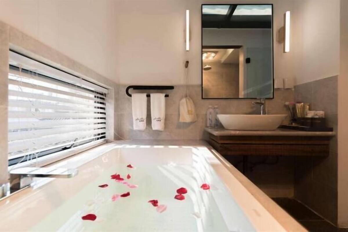 A modern bathroom with a large bathtub, a mirror, and a window with blinds.