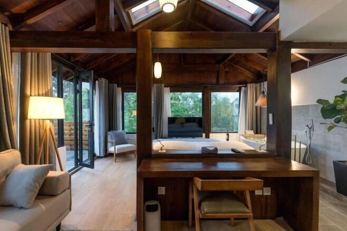 A modern bedroom with a large bed, a desk, and a chair. There is a window with curtains and a view of greenery outside.