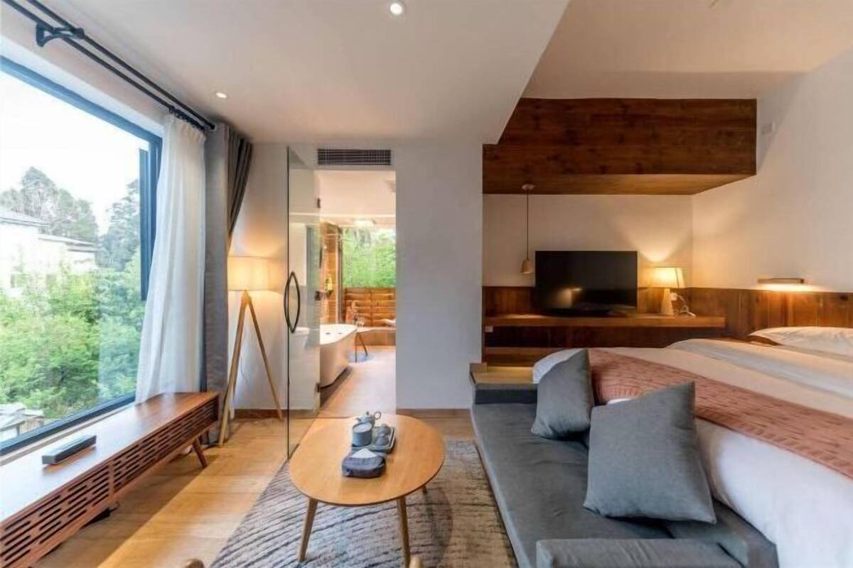A modern hotel room with a large window, a sofa, a bed, a TV, and a wooden ceiling.