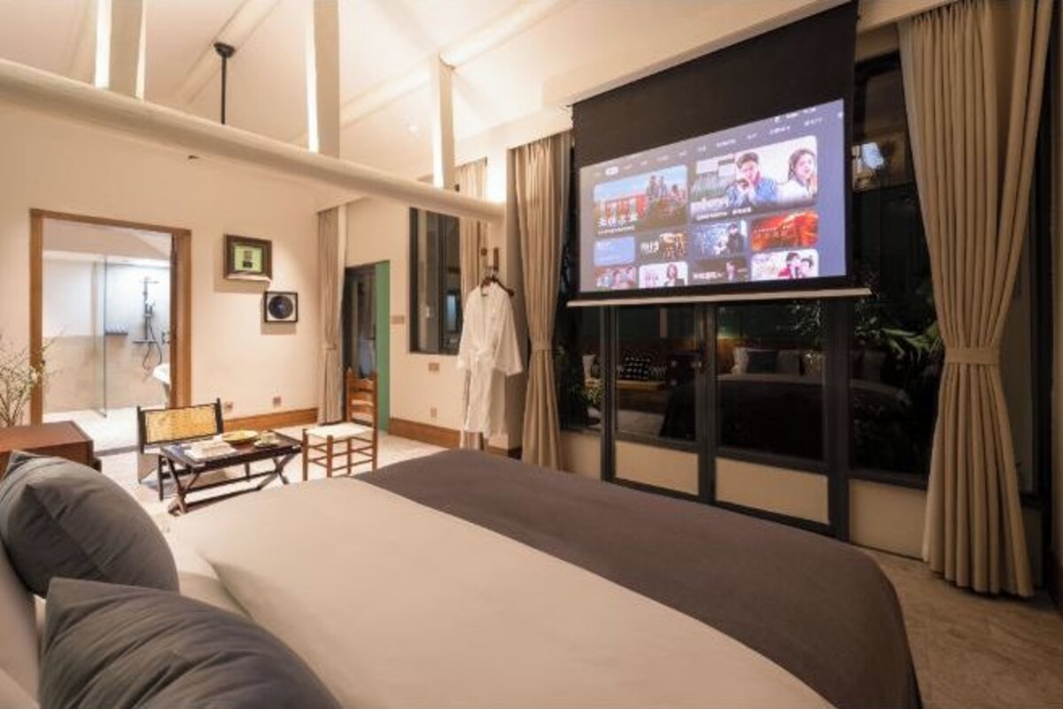 A hotel room with a large bed, a TV screen displaying a video conference, a seating area with a table and chairs, and a view of the outdoors through a window.