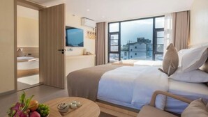 Une chambre d’hôtel moderne avec un grand lit, une table d’appoint en bois sur laquelle sont disposés des fruits, et une vue sur les bâtiments à travers de grandes fenêtres.