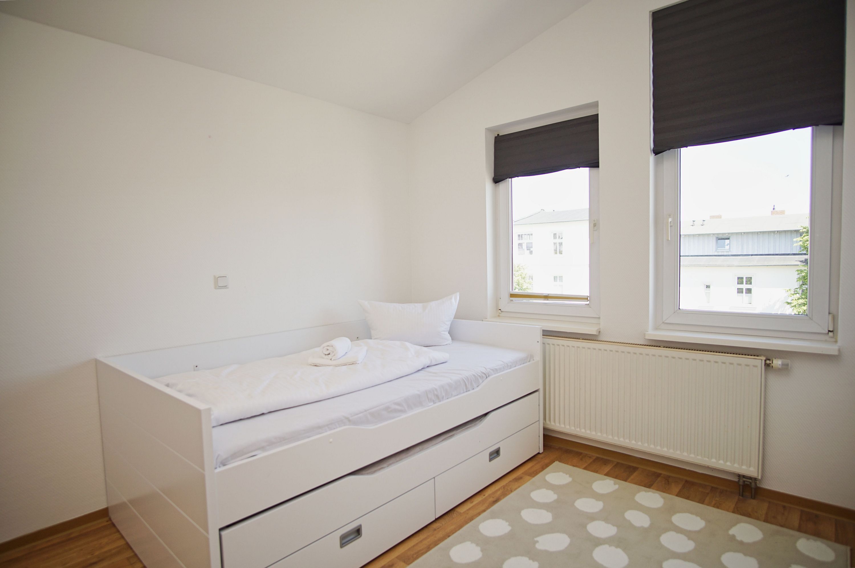A small, white bedroom with a single bed, a radiator, and two windows with dark blinds.