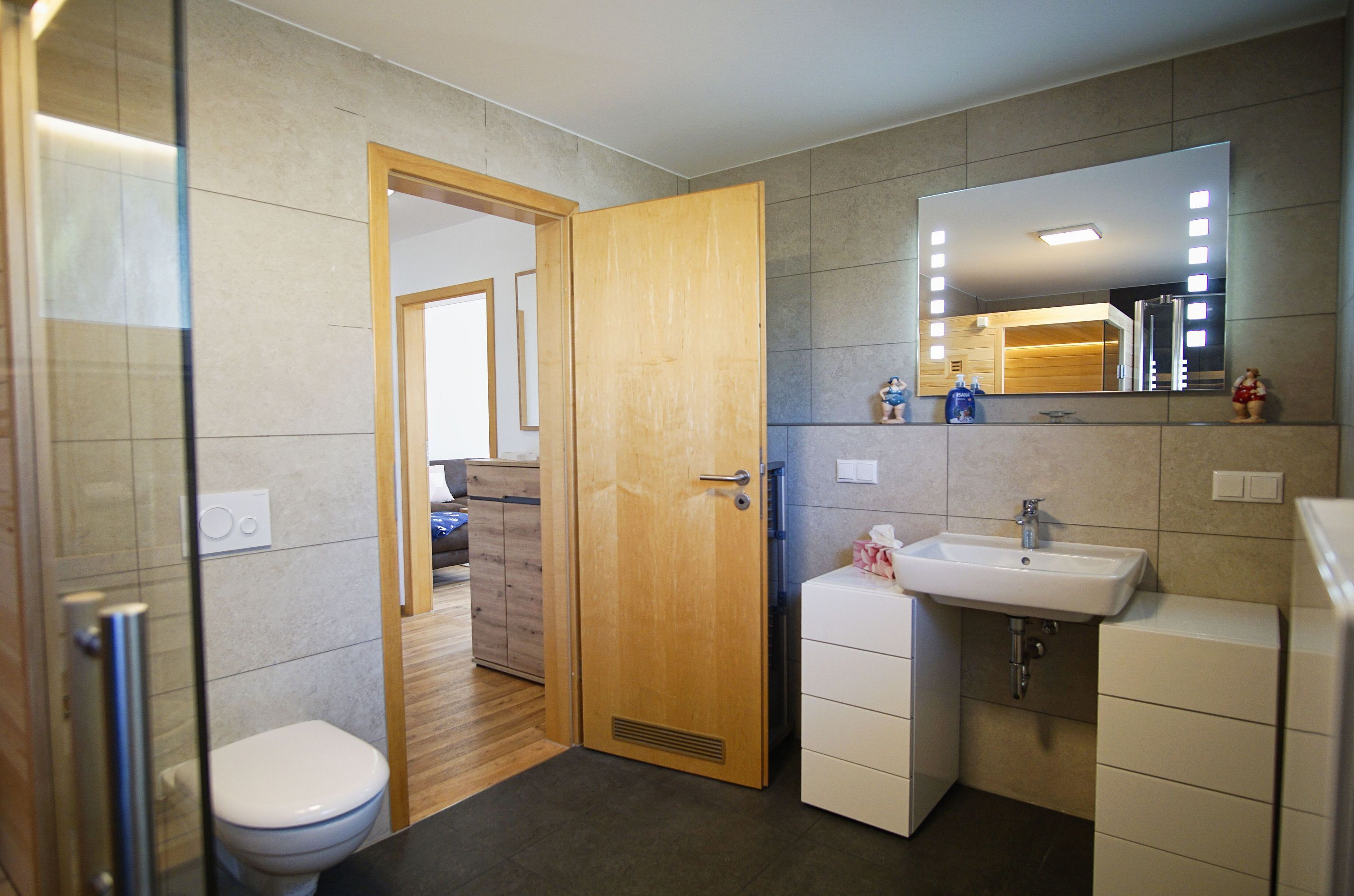 Une salle de bain avec une porte en bois, un lavabo blanc et un miroir avec éclairage intégré.
