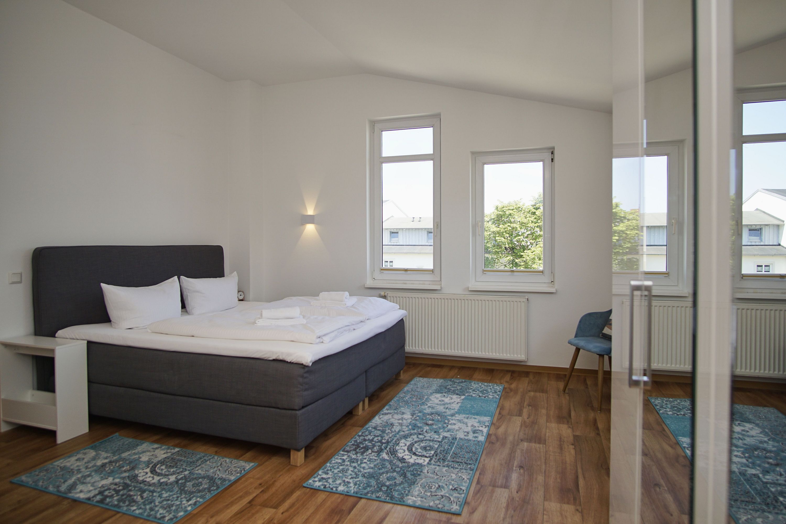 A bedroom with a bed, two windows, a chair, and a radiator.