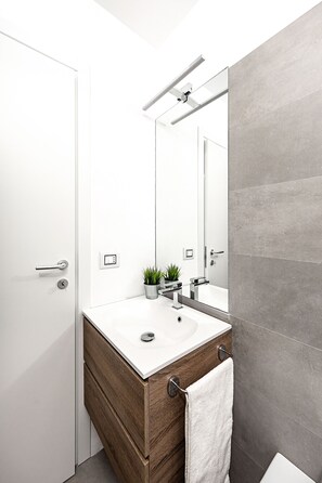 Un bagno moderno con un mobile lavabo bianco, uno specchio, una pianta in vaso e un asciugamano appeso alla porta.