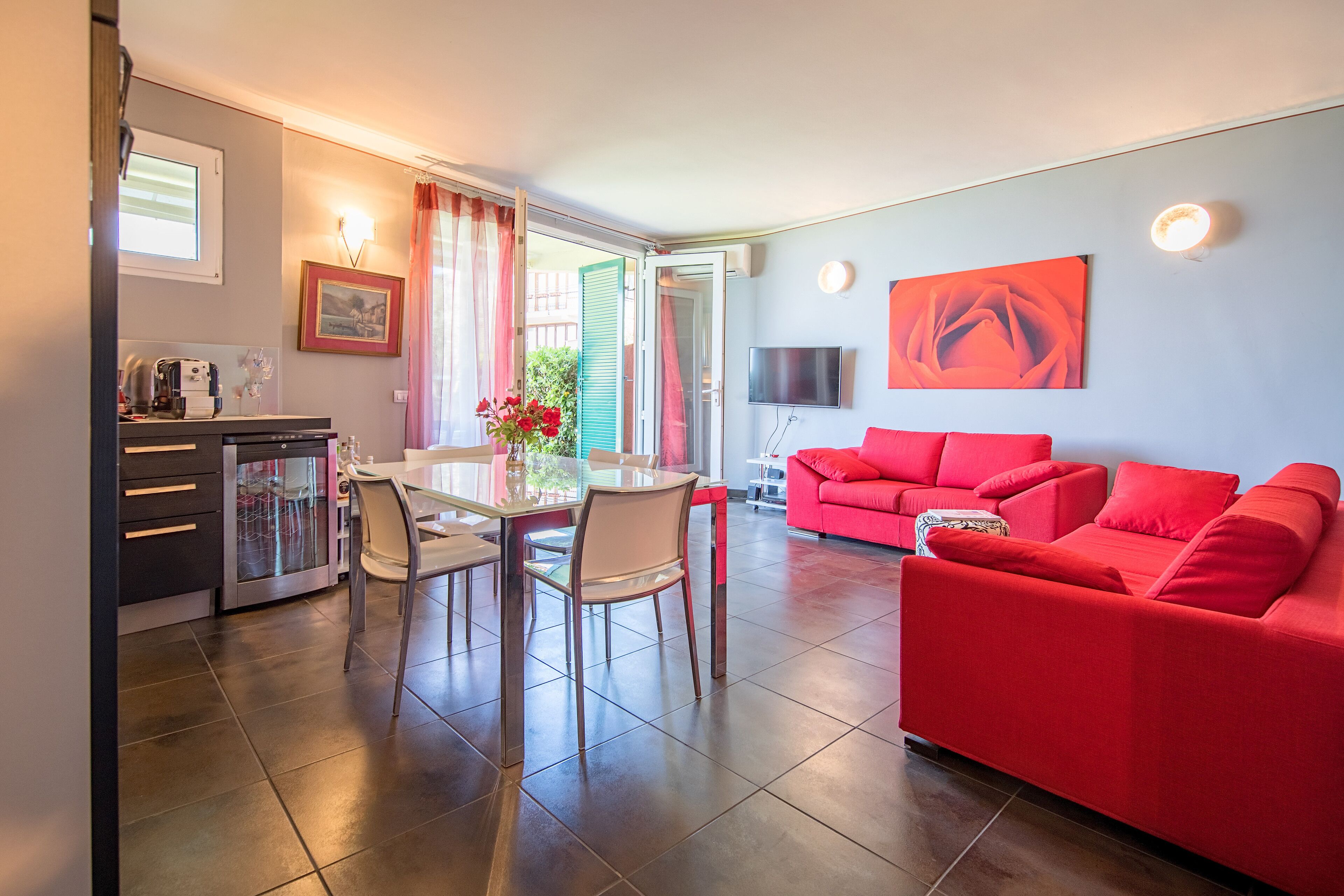 A modern living room with a red sofa, a dining area with a glass table, and a kitchen with a coffee machine.