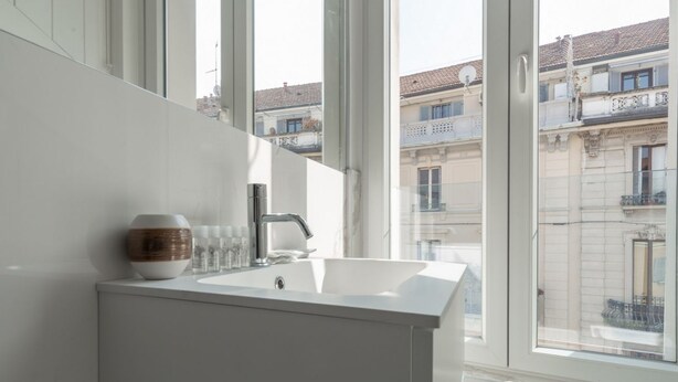 Ett modernt badrum med en vit tvättställ, en spegel och en dekorativ skål. Det har stora fönster som ger utsikt över ett bostadsområde med byggnader och balkonger.