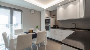 Ett modernt kök med vita skåp, en svart bänkskiva och en inbyggd ugn. Det finns en matplats med ett vitt bord och stolar, och en liten växt på bordet.