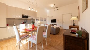 A modern kitchen with white cabinets, a dining area with a wooden table and chairs, a grey sofa, and a TV in the living room. - The Best Rent - Apartment near Isola Tiberina (Roma)