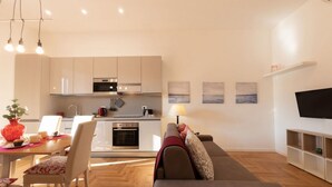 A modern kitchen with white cabinets, a built-in oven, and a microwave. There is a dining table with chairs and a red vase with flowers. A sofa and a TV are visible in the living area. - The Best Rent - Apartment near Isola Tiberina (Roma)