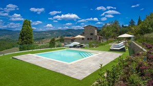 Un espace piscine doté d’une terrasse en pierre, de chaises longues et de parasols, entouré d’une végétation luxuriante et avec une chaîne de montagnes en toile de fond.