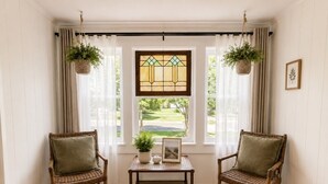 Quarto com duas cadeiras de vime, uma mesinha com planta e moldura, uma janela de vitrais e vista para uma paisagem verde.