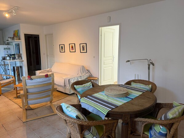 A cozy room with a bed, a dining table with chairs, a wicker chair, a standing lamp, and a door.