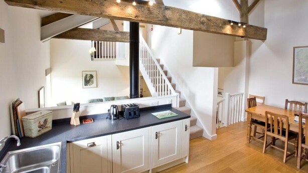 A kitchen with a wooden beam ceiling, a black countertop, white cabinets, a sink, and a dining area with a wooden table and chairs. - Nethergill Haylofts (Skipton)